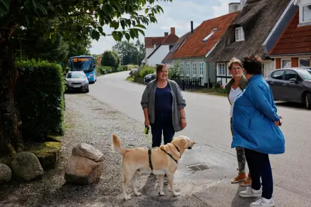 3 personer i samtale sammen med hund og lokalbus i baggrunden